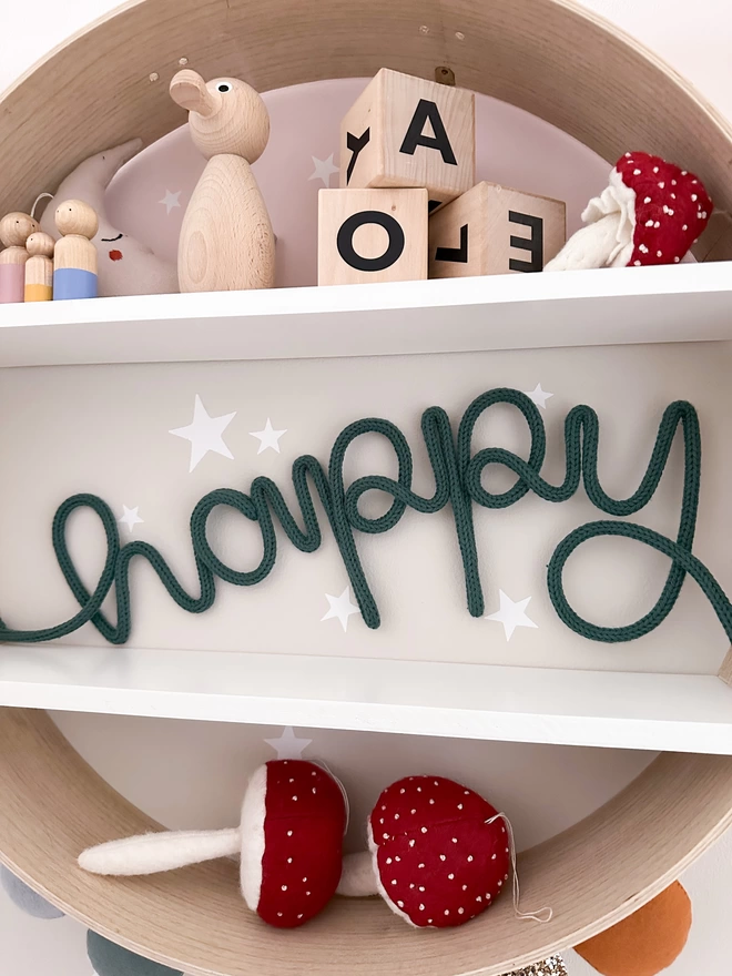 'happy' knitted wire word sign in a forest green colour on a circular book shelf decorated with mushroom toy props, wooden alphabet letter blocks and wooden duck toy. 