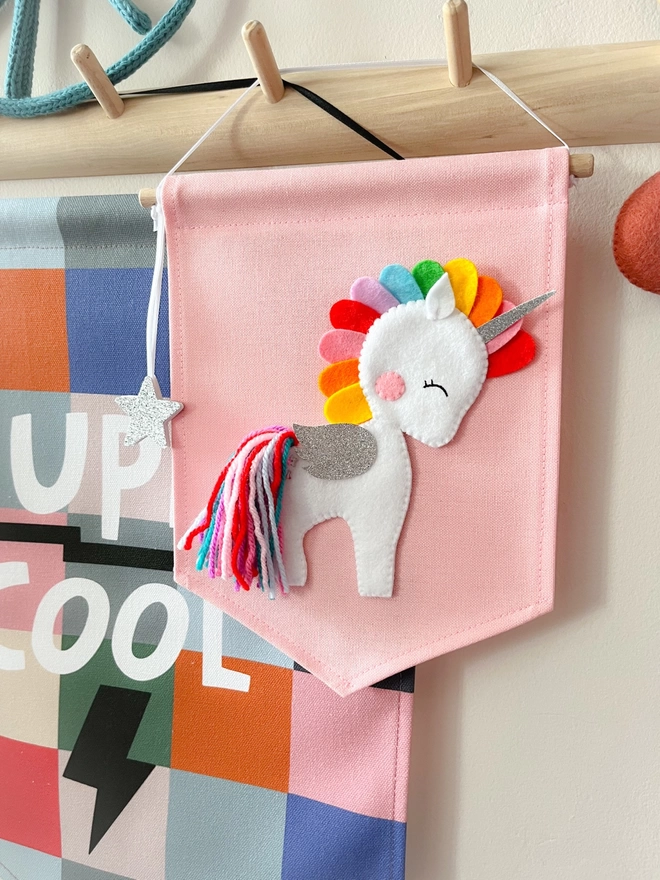 A white unicorn with a bright coloured mane and tail on a pink fabric banner hung from a peg rail