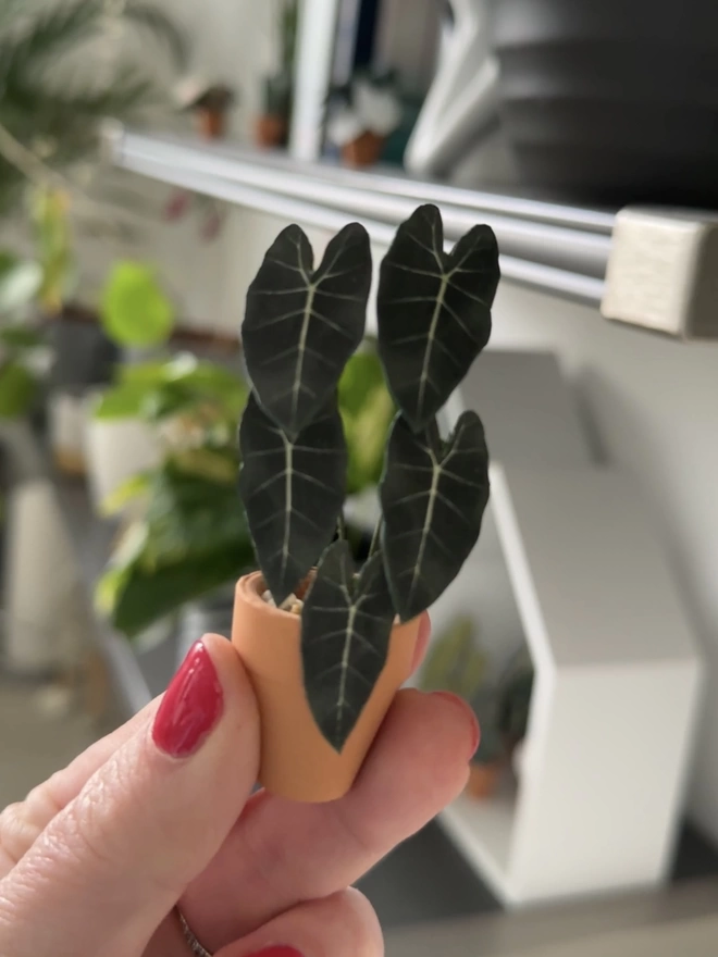 A miniature paper Alocasia Frydek being held between two fingers against a background of real plants