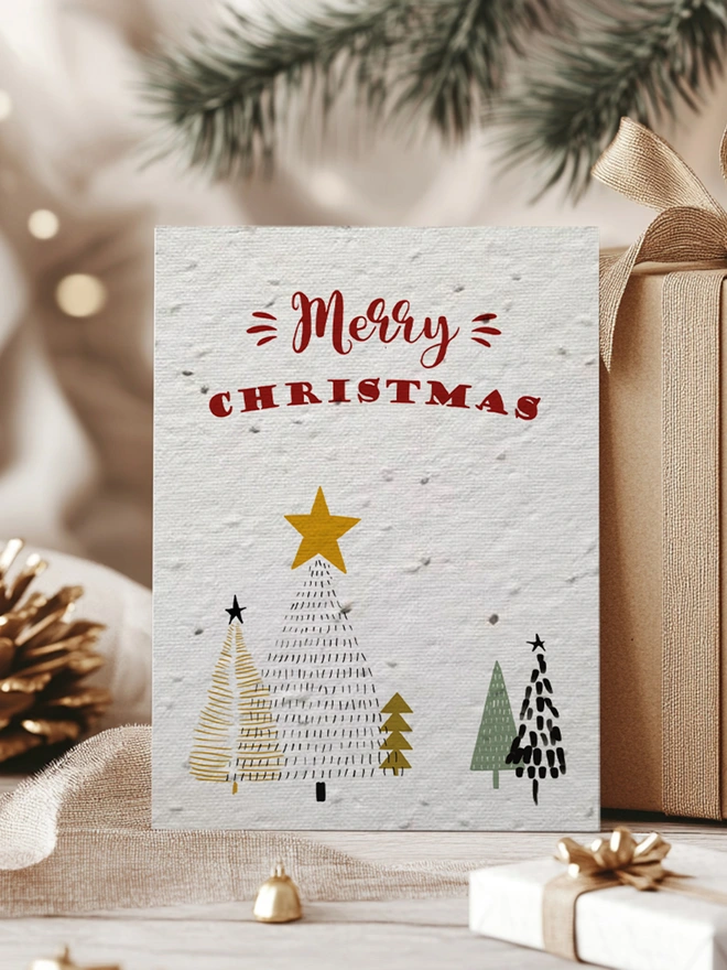 Christmas Card featuring Christmas trees and 'Merry Christmas' across the top. In the background is presents, pine cones and greenery.