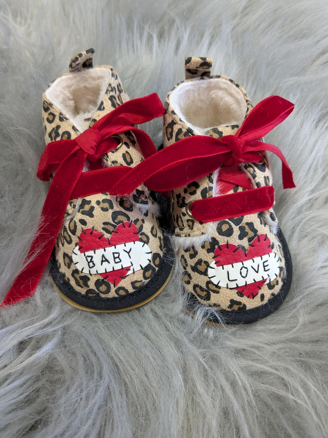 A pair of leopard print baby booties, lined with fur and with red velvet ribbon laces. A red heart with a white scroll adorns each booty. One says BABY, the other, LOVE in black stitches
