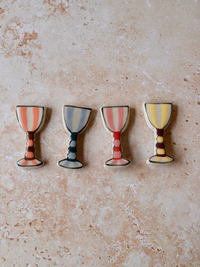 Handmade ceramic magnets shaped like small goblets, featuring colourful stripe designs in orange, blue, pink and yellow, aligned on a neutral stone surface.