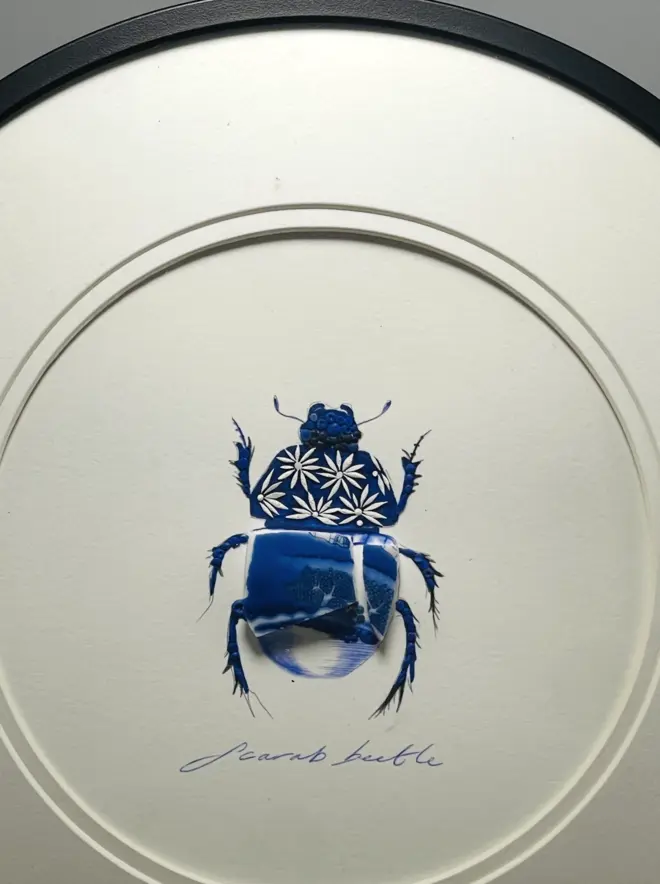 Hand-drawn, painted, mosaic clay, vintage Willow Tree pottery insect on white double mounted card, in a black circular frame.