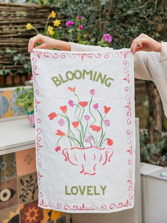 Linen teatowel with Blooming Lovely and floral pattern, held up against a garden background