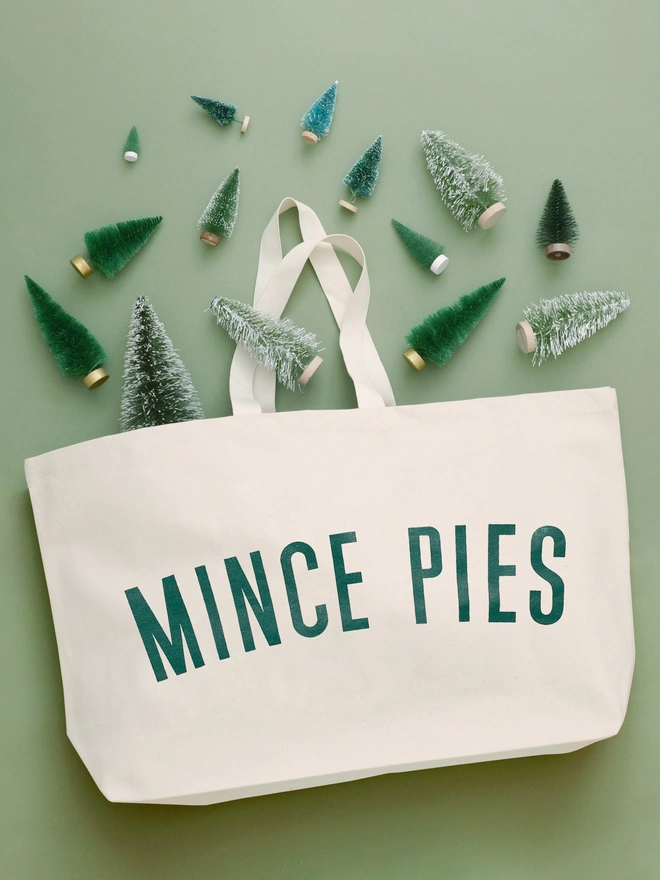 an oversized natural canvas tote bag with bold mince pies slogan laying on a green backdrop with mini christmas trees surrounding