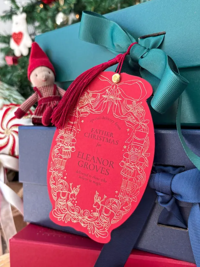 A personalised red gift tag with a burgundy tassel, tied to a teal gift box with a green grosgrain bow. Its gold foil border shows nutcrackers, reindeer, snowflakes and a ribbon bow, around the words "Special delivery from Father Christmas for Eleanor Groves, delivered to those who believe in magic". A small elf doll and a Christmas tree sit behind it.