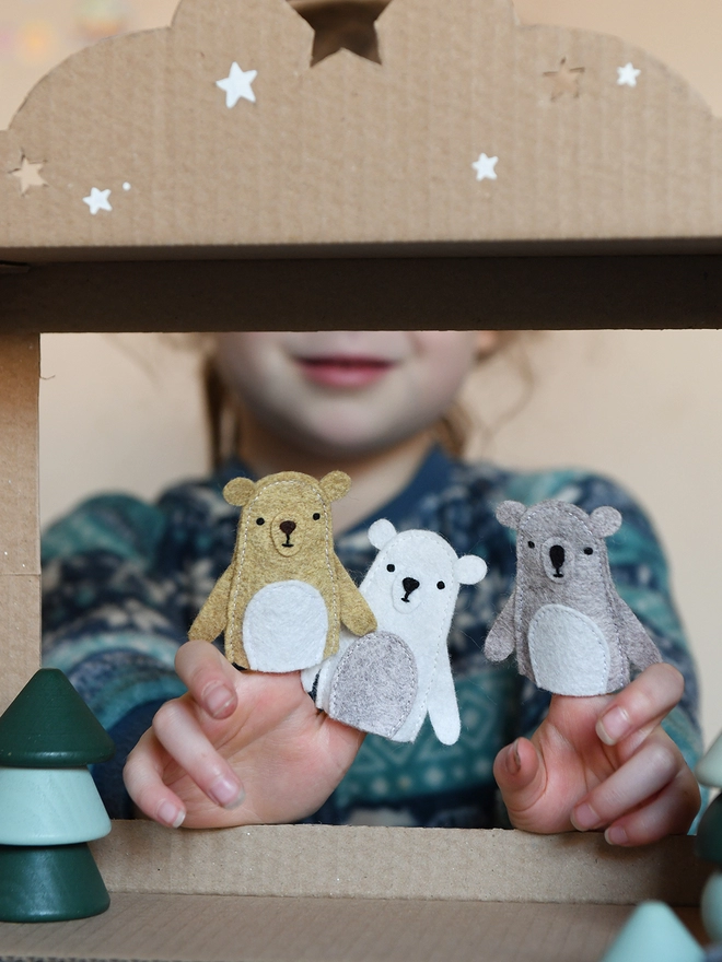 A young child wearing a blue jumper holds three handmade felt bear finger puppets on their fingers.