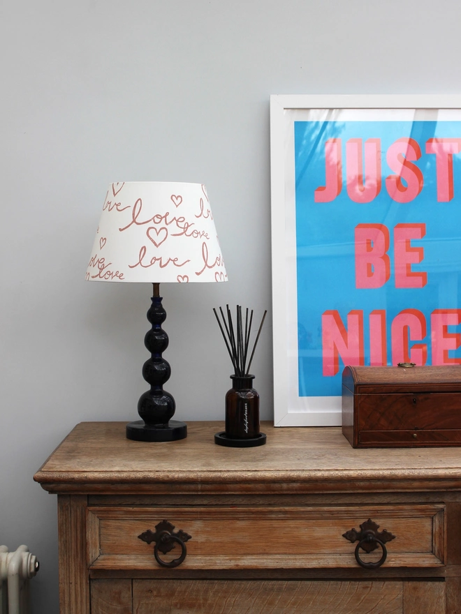 Studio Zef hand-painted lampshade with playful pink ‘Love’ lettering and hearts, styled on a blue table lamp against a modern interior with vintage wooden dresser and bold wall art.