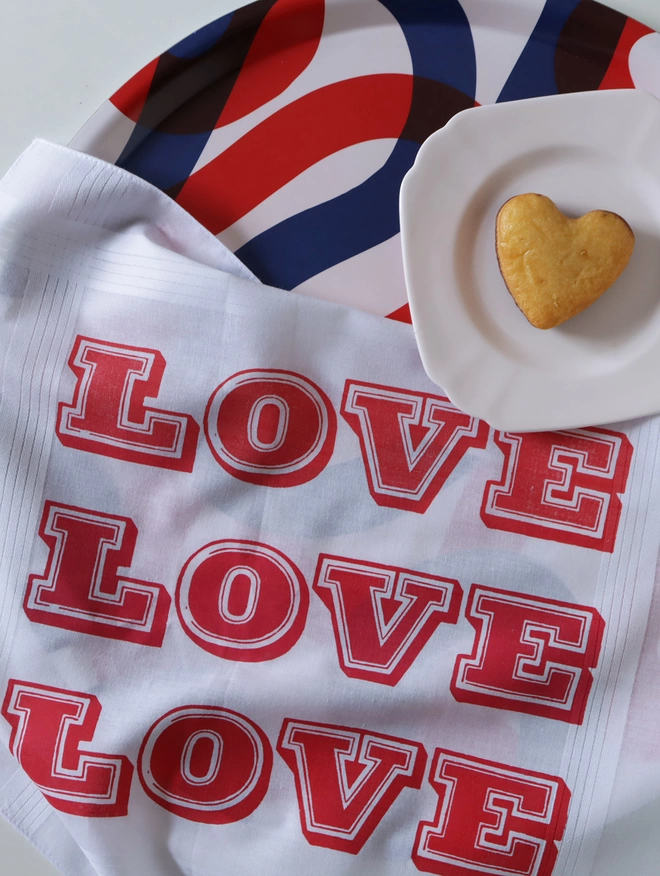A Mr.PS LOVE LOVE LOVE handkerchief alongside a heart shaped cake on a tea tray