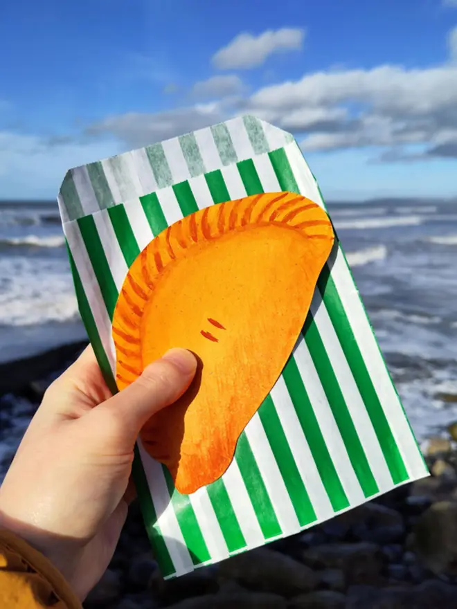 hand painted cornish pasty drawing, with green and white striped paper bag, held in front of ocean view