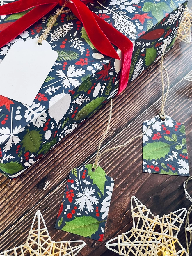 Close-up of present wrapped in Winter leaf wrapping paper with Holly and Ivy printed design, red bow, and matching gift tags on wooden background with star lights