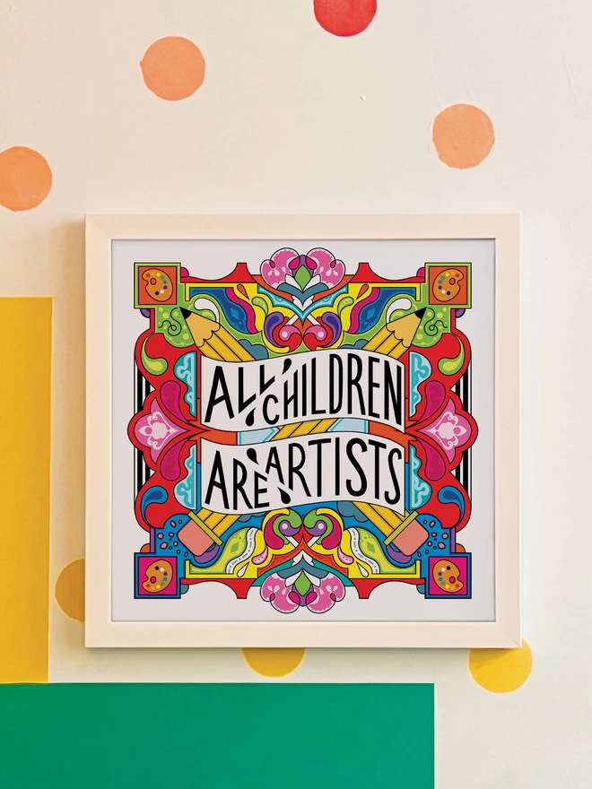 A white square frame surrounds the print, which has the words All Children are Artist at the centre of this multi-coloured vibrant illustration which includes a cross of pencils, and artists palettes the corners. It is hung on a white wall with yellow, orange, green and blue spots, and a green and yellow rectangle painted in the bottom left hand corner.