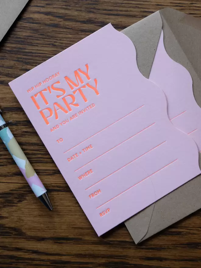 Wavy cut pink party invite, letterpress printed with bright orange ink. Set out on a table with kraft envelope.