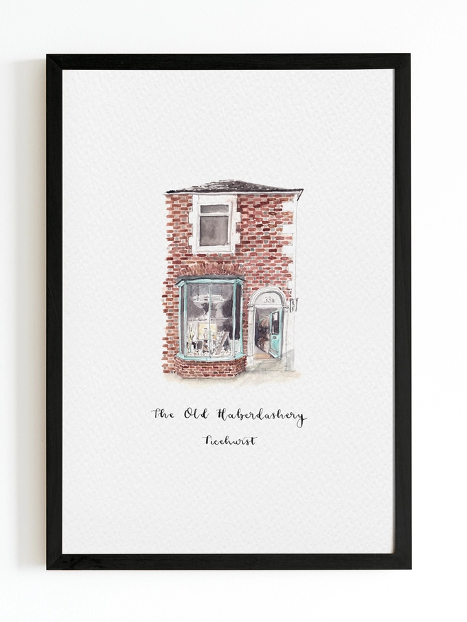 Watercolour painting of The Old Haberdashery, haberdashery and gift shop in Ticehurst, a beautiful brick building with light blue framed window and door which is open welcoming you into the shop. The watercolour style is painted with a black pen outline and organic loose style with small details. The illustration sits on a white piece of paper with a black frame.
