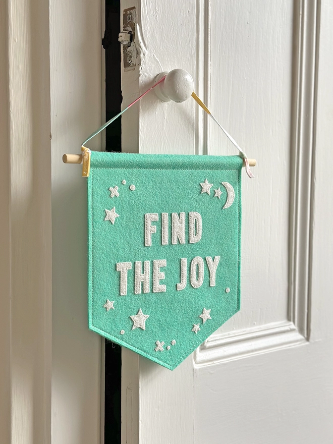 A turquoise felt banner with white words that read "Find The Joy" stitched on hangs from a ribbon hanger on a white wooden door.
