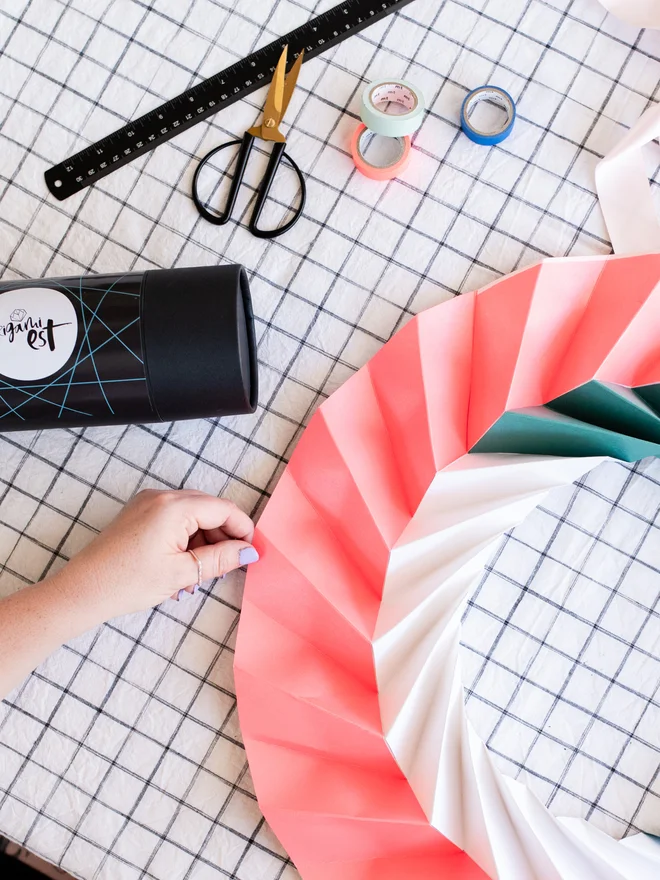 Woman holding an origami kit in a black tube with paper ornament and craft materials in background.