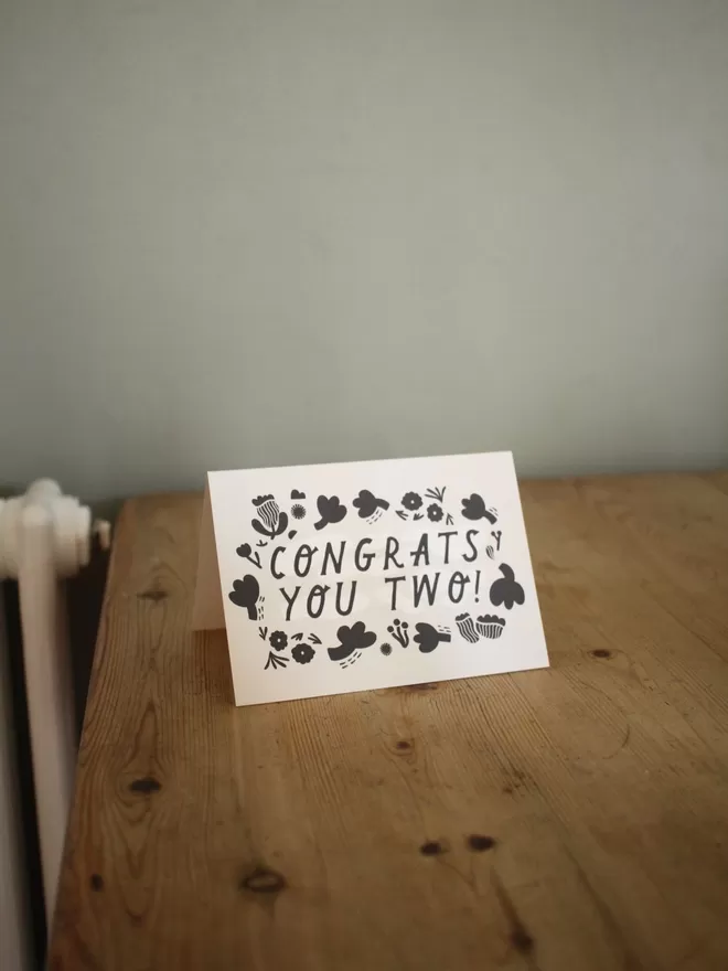 Black and white greeting card with illustration and the words Congrats you two written on it stood up on wooden surface 