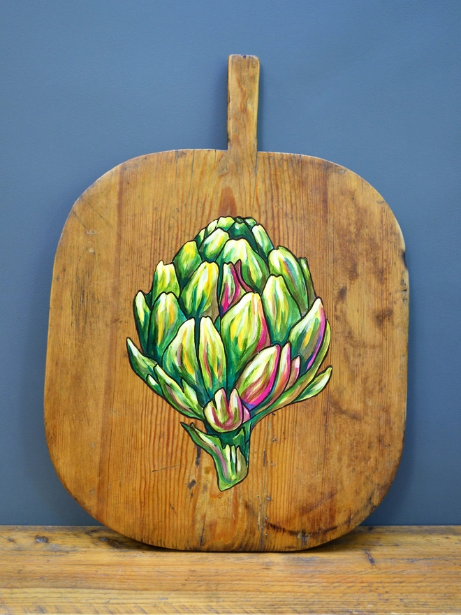 Wooden chopping board with handpainted design of an artichoke standing against a wall