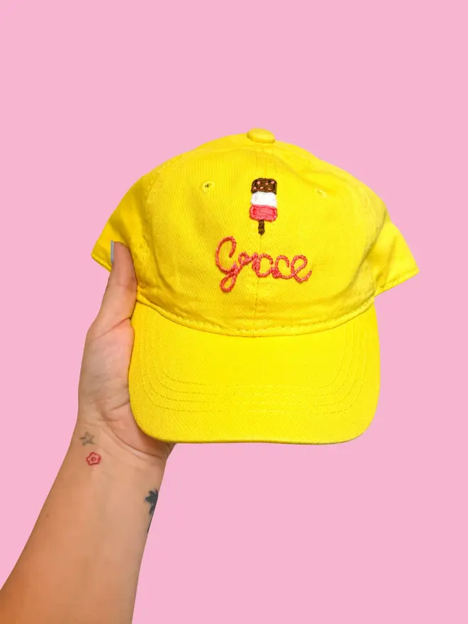 Hand Embroidered Kids Cap, a person holding a personalised cap against a colourful backdrop. It says a child's name. 