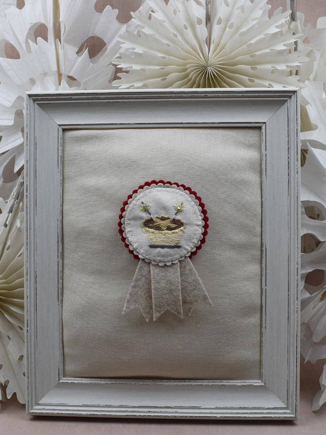 Christmas red and Beige felt rosette with embroidered Mince Pie as the central design.  Pinned to a padded frame with paper snowflakes in the background.