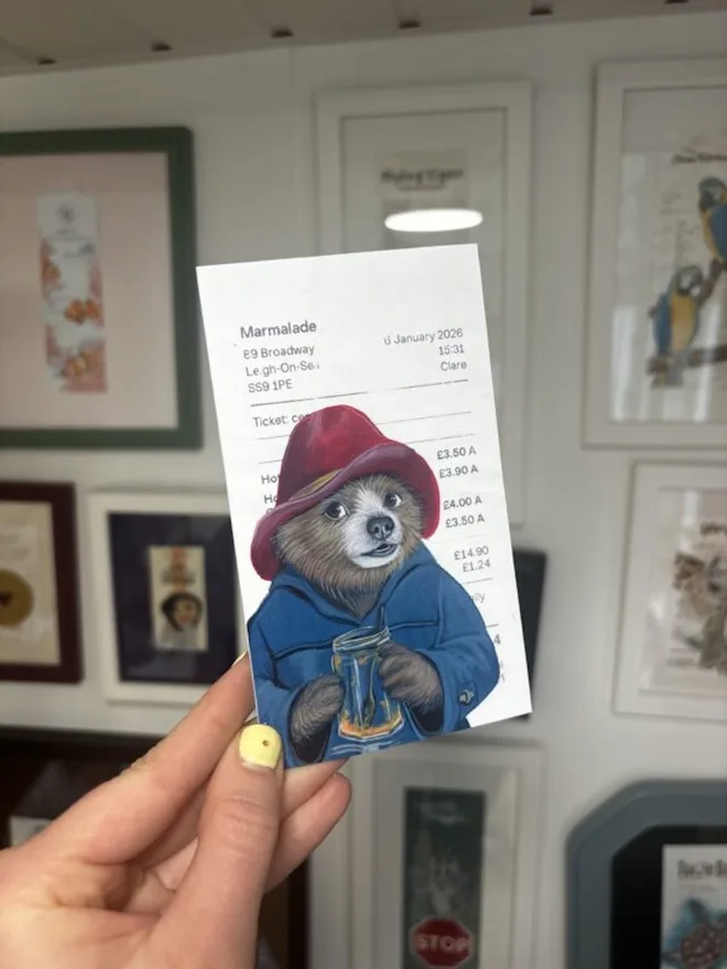 Out Of Marmalade Paddington Print, being held in a room with drawings. 