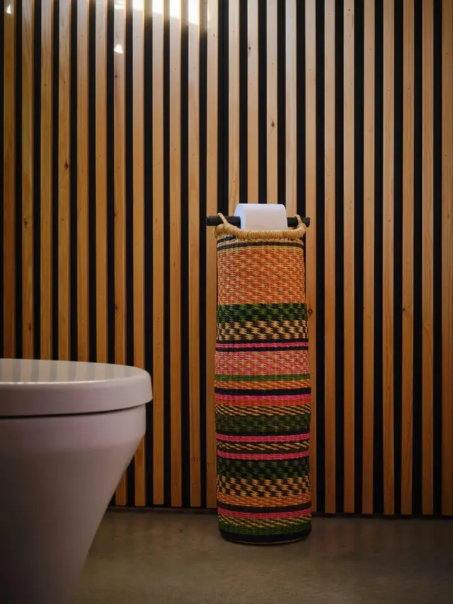 Ghanaian Hand Woven Tubular Toilet Roll Holder, a toilet roll holder sitting in a bathroom. It sits in front of a wooden backdrop. 