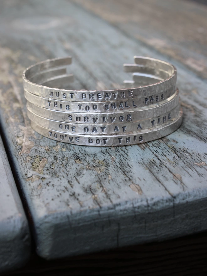 A selection of sterling silver cuff bangles with stamped text on, stacked together on a weathered blue painted table.