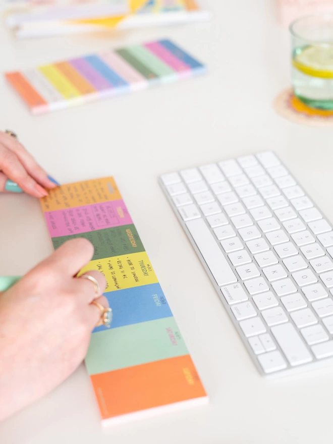 A woman writes on the colourful Raspberry Blossom Keyboard Jot Pad, a keyboard is above the jot pad with a glass of water and other items from the Happiness Stationery Collection