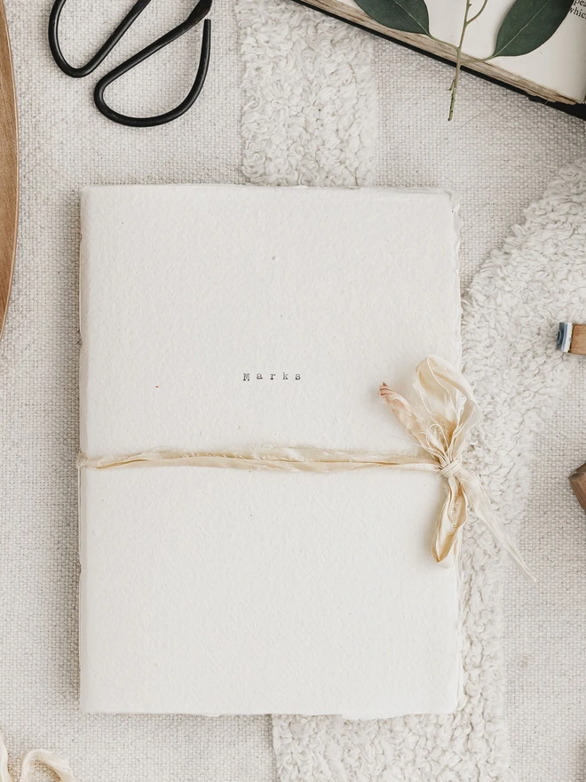 an off white journal with a deckled edge and rustic ribbon closure the word 'marks' has been typed on the cover
