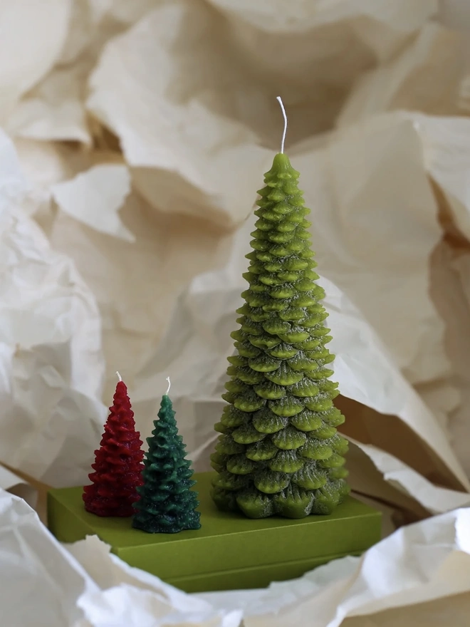 Large Christmas Tree Candle. a large lime green christmas tree shaped candle stands on a green box, nestled among crumpled cream tissue paper. a smaller red and a smaller dark green tree candle sit in front of it for scale, both with visible wicks. the textured, layered pine branch design and slightly variegated wax give all three candles a realistic, hand-poured finish.