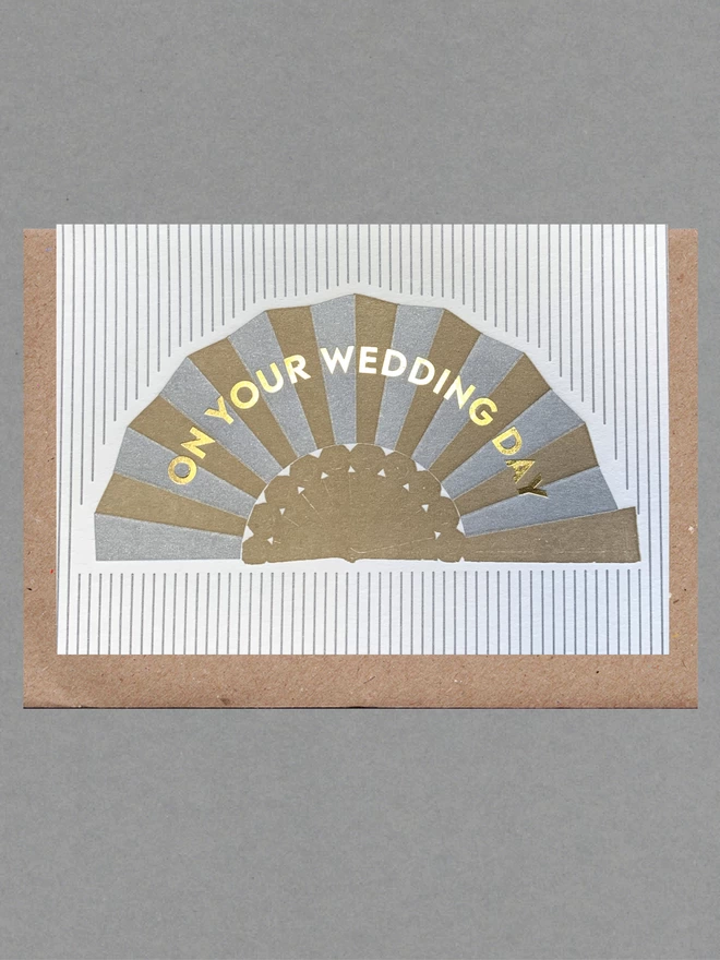 White and silver striped card with silver and gold fan on it with  gold text reading 'On Your Wedding Day' on it with a brown envelope behind