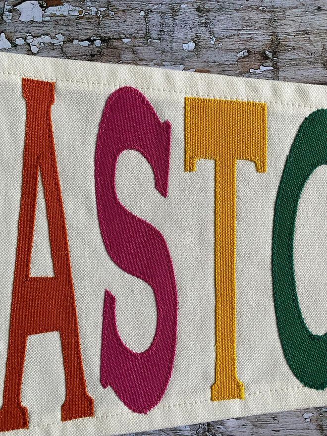 Detail of an ivory canvas Glastonbury pennant flag showing the letter ASTO inn orange, pink, yellow and green canvas.
