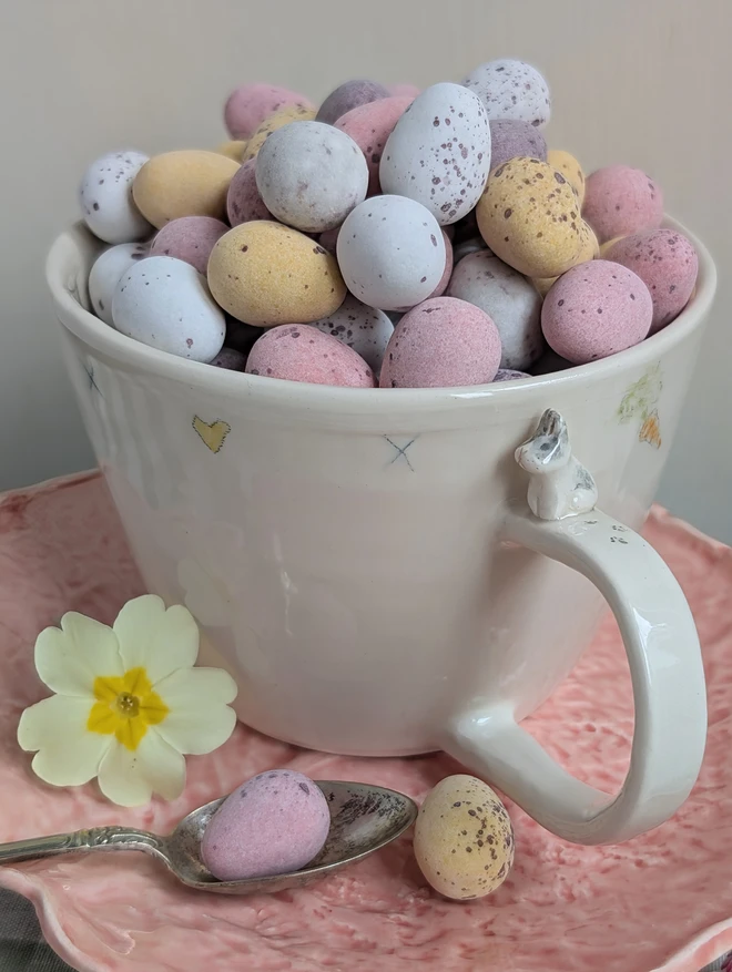 Bunny rabbit cup with tiny rabbit on the handle with pawprints hearts kisses and carrots illustrated on the mug that is filled with Easter mini eggs