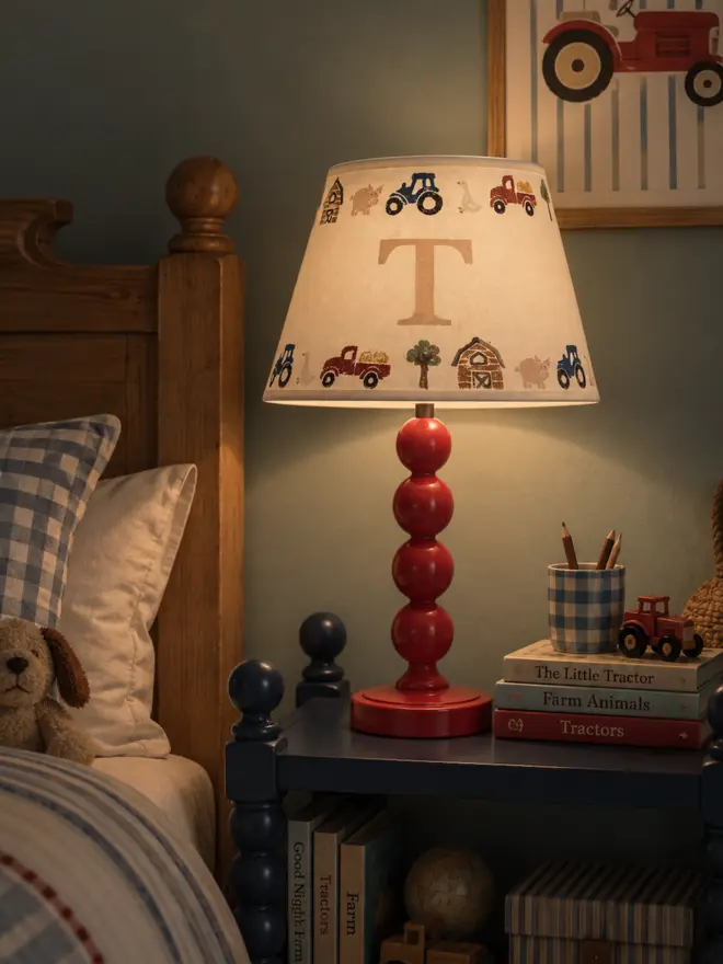 Glowing personalised children's farmyard lampshade by Studio Zef, featuring hand-printed blue tractors, red farm trucks, pigs and barns surrounding a large initial T. The cream empire lampshade sits on a glossy burgundy red bobbin lamp, casting a warm, cosy light across a traditional boy's bedroom with gingham bedding, wooden furniture and farm-themed accessories.