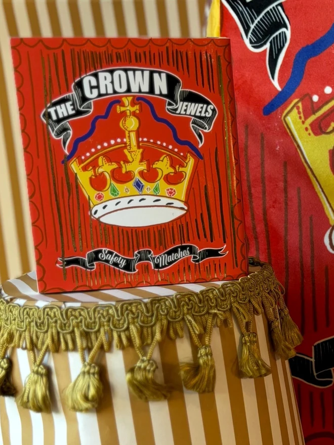 The Crown Jewels Matchbox, a red matchbox with a crown design sitting on a decorated surface accompanied by other accessories. 