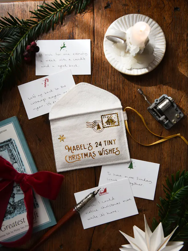 A handmade felt envelope advent calendar with golden lettering that reads 'Mabel's 24 Tiny Christmas Wishes' with several Christmas notecards all laying on a wooden table.
