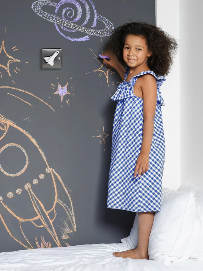 A brushed chrome dimmer light switch with a space shuttle as the rotary knob to turn the lights on and off on a chalk board style bedroom wall. A six year old black girl in a blue and white dress stands on a bed with a white pillow and white duvet, she faces the camera smiling as she draws on the wall in chalk. The children's light switch brand is Candy Queen Designs.