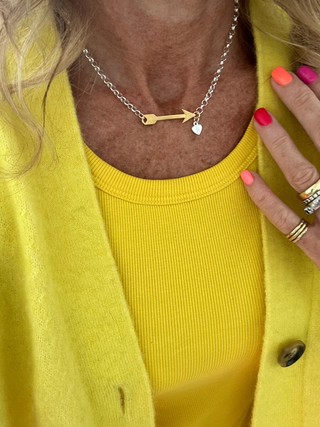 model wears a gold arrow charm linked at either end to a sterling silver chain, a small silver heart charm sits at the point of the arrow