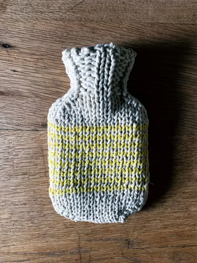 Mini Hot Water Bottle in hand knit string cover with yellow stripe on an oak table