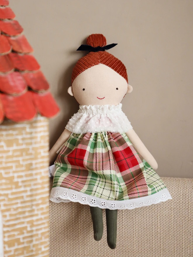 Handmade cloth doll with red brown corduroy hair in a top knot tied with a navy velvet bow, wearing a white textured ruffle collar, a red and green tartan dress with lace trim, and dark green tights.