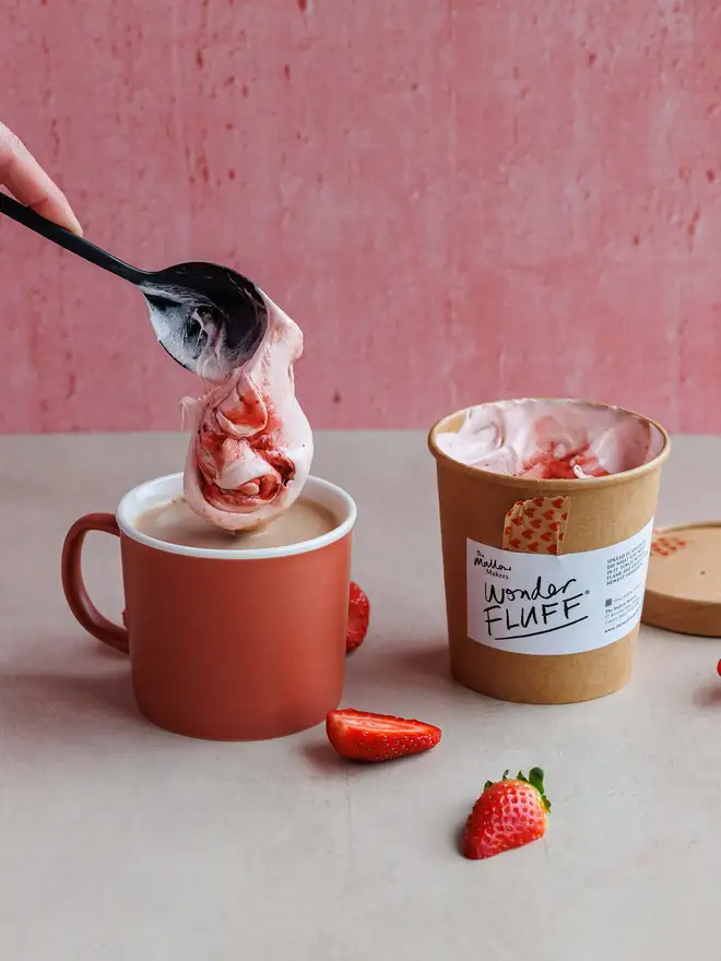 Strawberry Wonder Fluff Tub, a hot chocolate with strawberry fluff being dropped on top from a spoon. This is on a white surface with an opened tub in the background and fresh strawberries scattered around the bottom.