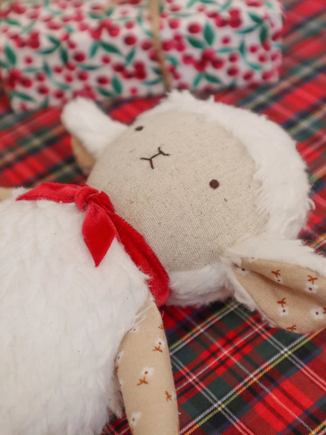 Handmade lamb doll with a fluffy white body, stitched face, beige floral fabric arms and legs, and a small red velvet bow tied around its neck.