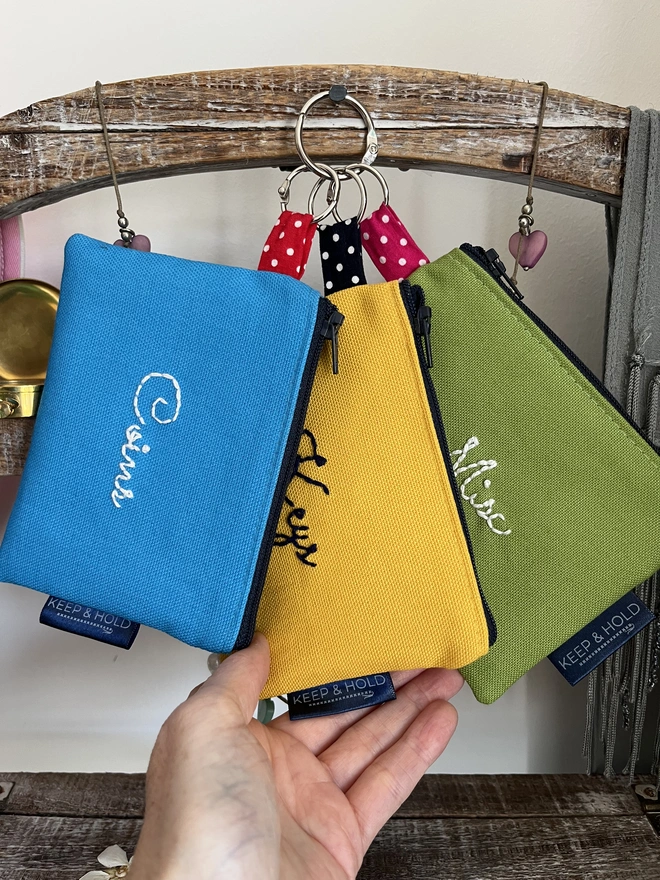 3 zipped purses in blue, yellow and green, embroidered on the front. Hanging from a wooden stand.