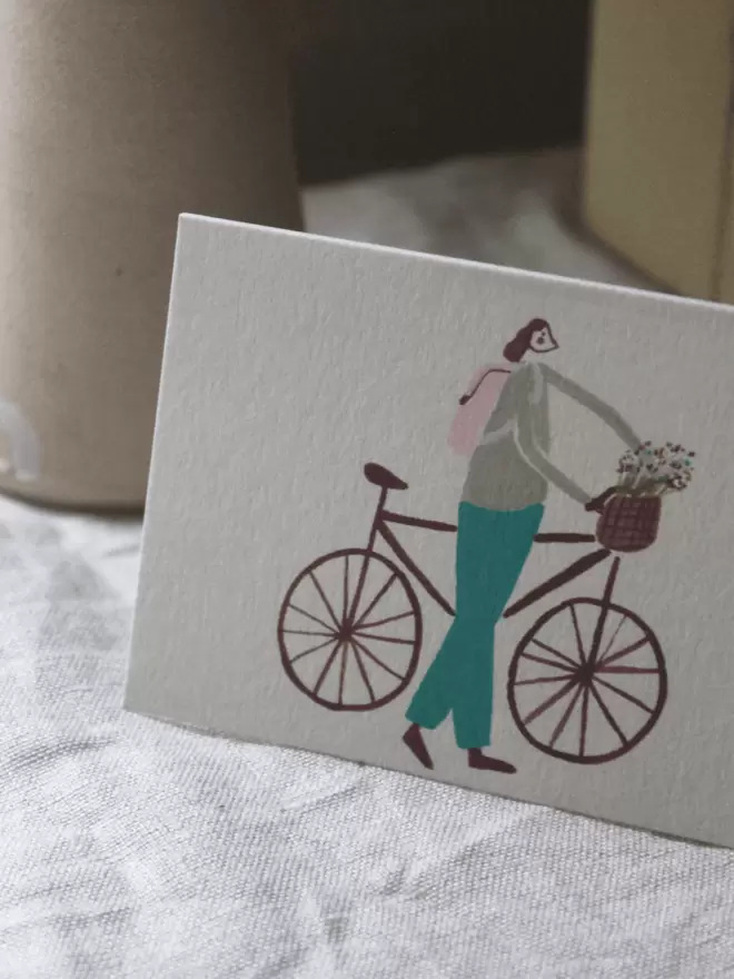 a greetings card with a girl riding a bike with flowers in the basket. 