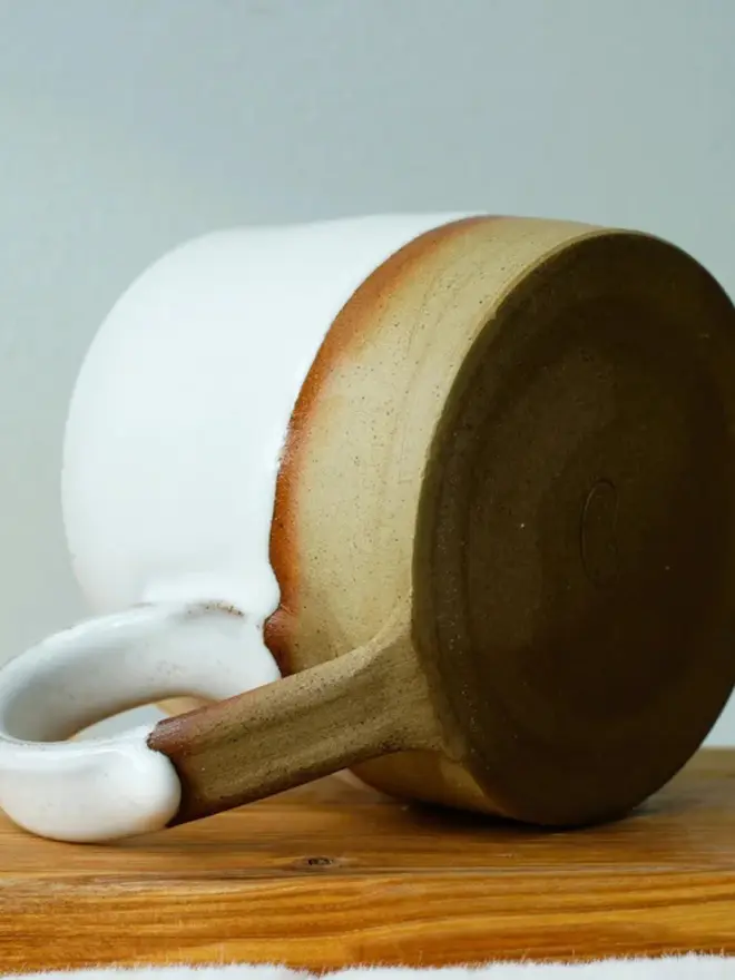 Handmade ceramic mug with a glossy white upper glaze and a natural toasted clay base, resting on a wooden surface against a soft green wall background.