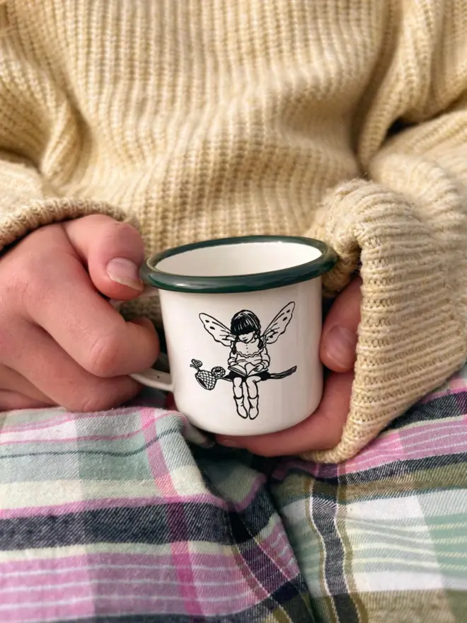 a small child holding a miniature enamel mug cream in colour with a bottle green rim featuring an illustration of forest folk on a branch