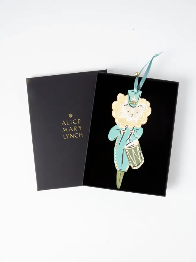 Lion Drummer Tree Dancer. wooden hanging lion drummer decoration in an open black gift box embossed "alice mary lynch" in gold, with duck egg blue tunic, cream mane and olive green drum, blue ribbon with a jingle bell