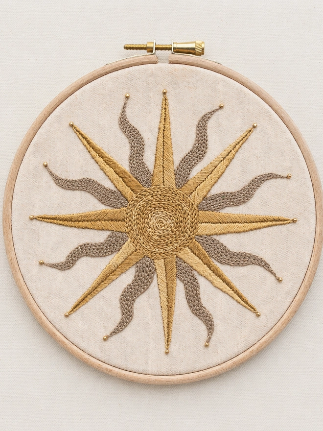 Completed Golden Solstice embroidery design featuring a celestial sun motif stitched in gold and taupe thread on natural cotton fabric in a wooden embroidery hoop.