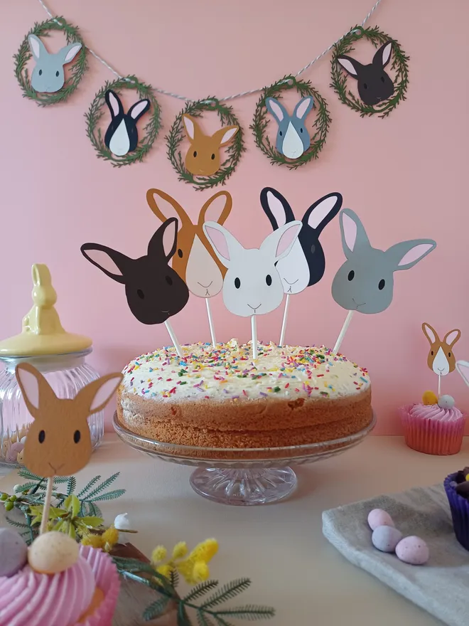 Easter Bunny Garland displayed on pink wall behind large cake with 5 Easter Bunny Cake Topper Decorations and small easter bunny cupcake toppers. Bunny decorations are grey, white, dark brown, black and white with pale pink ears