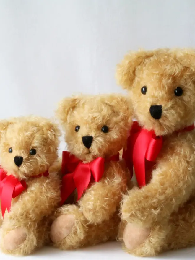 Three brown handmade teddy bears sitting side by side in a row, each wearing a red ribbon around their necks.